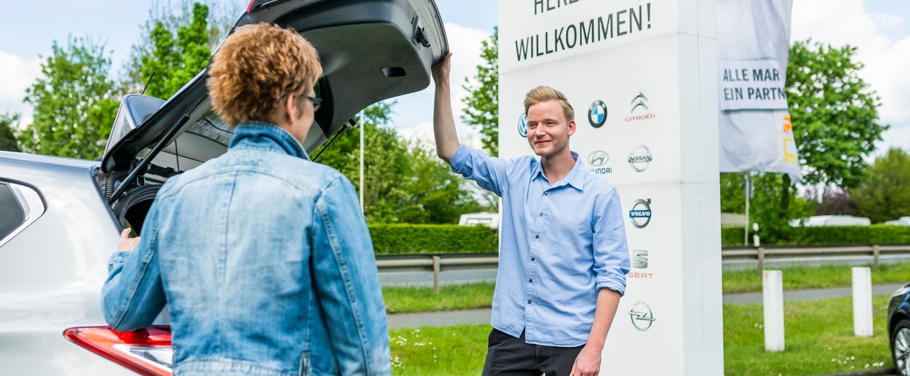 Ein Verkäufer im Dialog mit einer Kundin im Außenbereich an einem Auto mit offenem Kofferraum