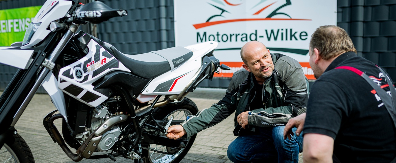 2 Männer in der Hocke schauen sich ein Motorrad an, im Hintergrund Werbewand Motorrad Wilke